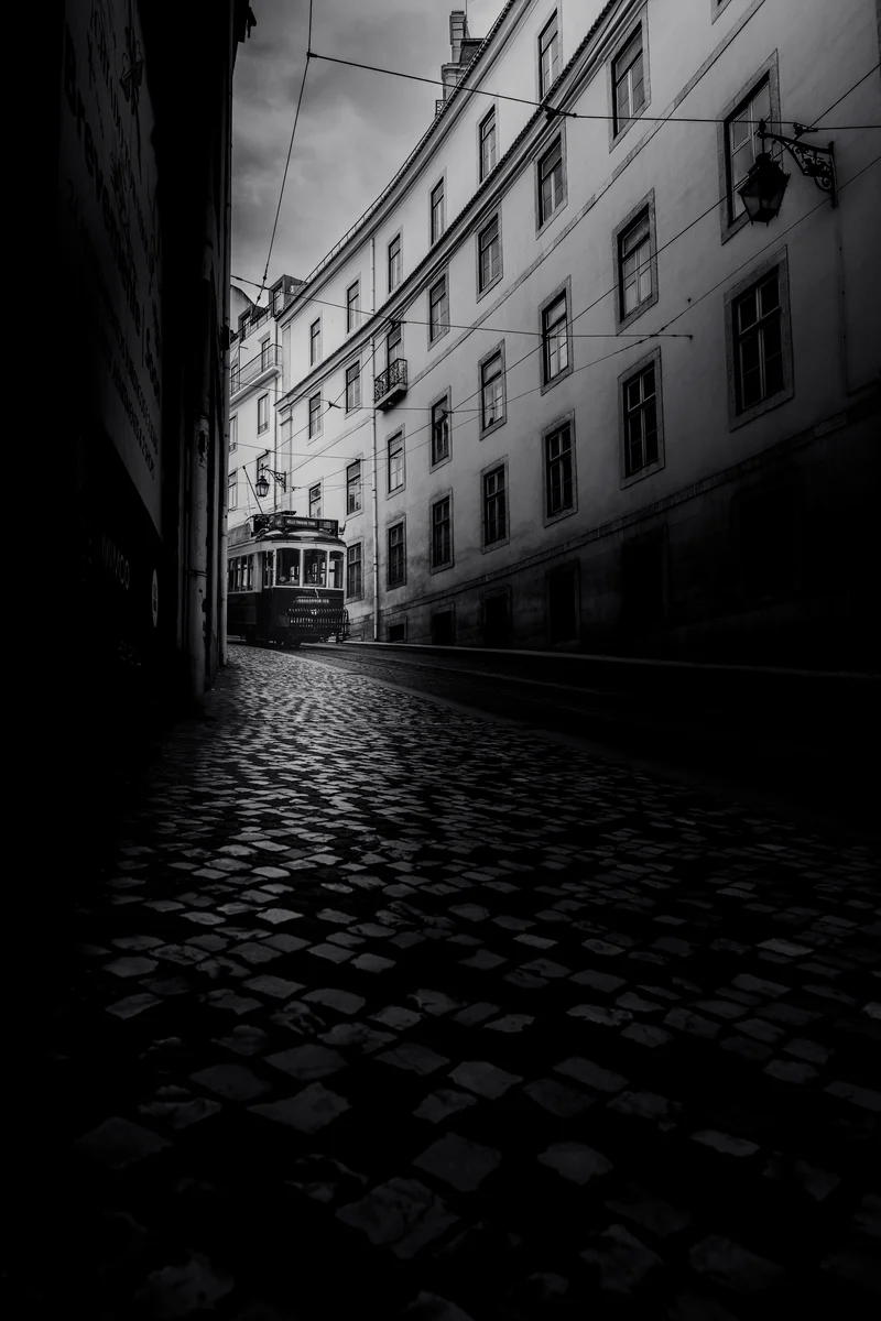 Narrow cobbled street with a tram emerging from shadow in black and white Porto scene (Mustvalge stseen Portost – kivisillutisega tänav ja tramm valgusest välja sõitmas)