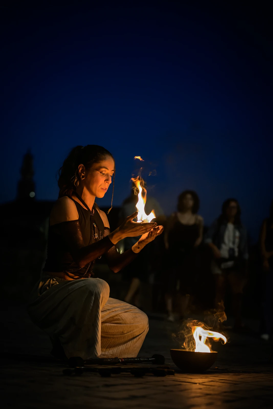 Girl holding a candle at night, face lit by flame, people sitting around her (Tüdruk istub küünlaga, öine tuli valgustab vaikselt)