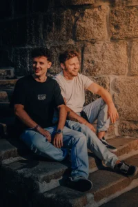 Two men sitting casually on stone ledge, golden light hitting textured wall (Sõbrad istuvad kiviseina ääres päikeseloojangus)