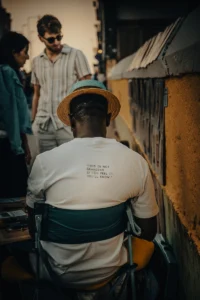 Street artist from behind wearing “Truth is not outside” shirt at art stand (Tänavakunstnik tagantvaates, kunstiseina ees istumas)