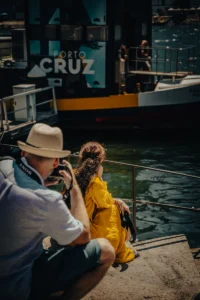 Photographer in hat capturing woman in yellow by riverside near “Porto Cruz” boat (Porto jõeäärne stseen, dokumentaalne hetk)