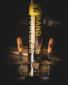 Lähiplaan läikivast nurgakivi kapslist kirjaga "RAND & TUULBERG" ja kahest graveeritud kellust puidust alusel.