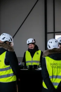 Naeratav ehitustööline Rand & Tuulberg logoga kiivris ja helkurvestis vestlemas kolleegidega siseruumis.