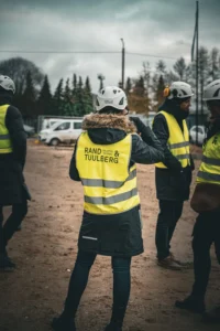 Naine Rand & Tuulberg logoga helkurvestis ja kiivris vaatamas seljaga kaamera poole ehitusplatsile.